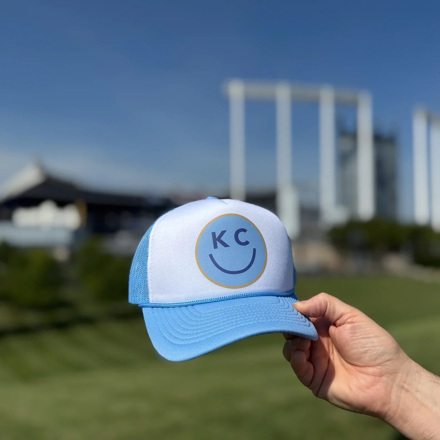 Blue and white KC Smile foam trucker hat with 'KC' logo held in front of a stadium.