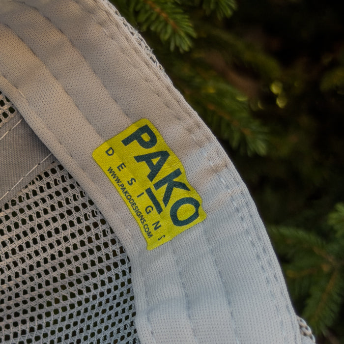 Close-up of a beige backpack with a yellow 'PAKO Designs' label against a green foliage background.
