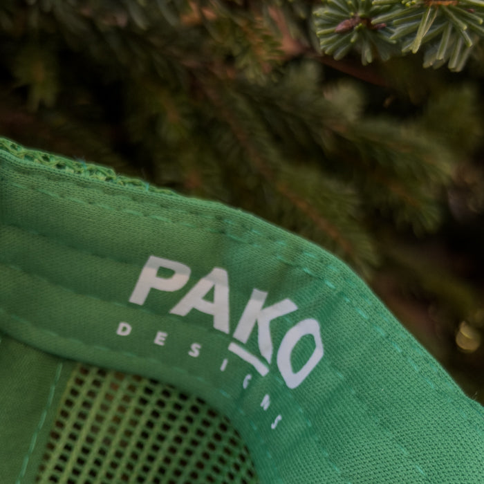 Green cap with 'PAKO DESIGNS' logo against a blurred natural background