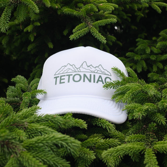 A PAKO Designs white 5 panel foam trucker hat with Tetoniac design on the front, displayed in a pine tree