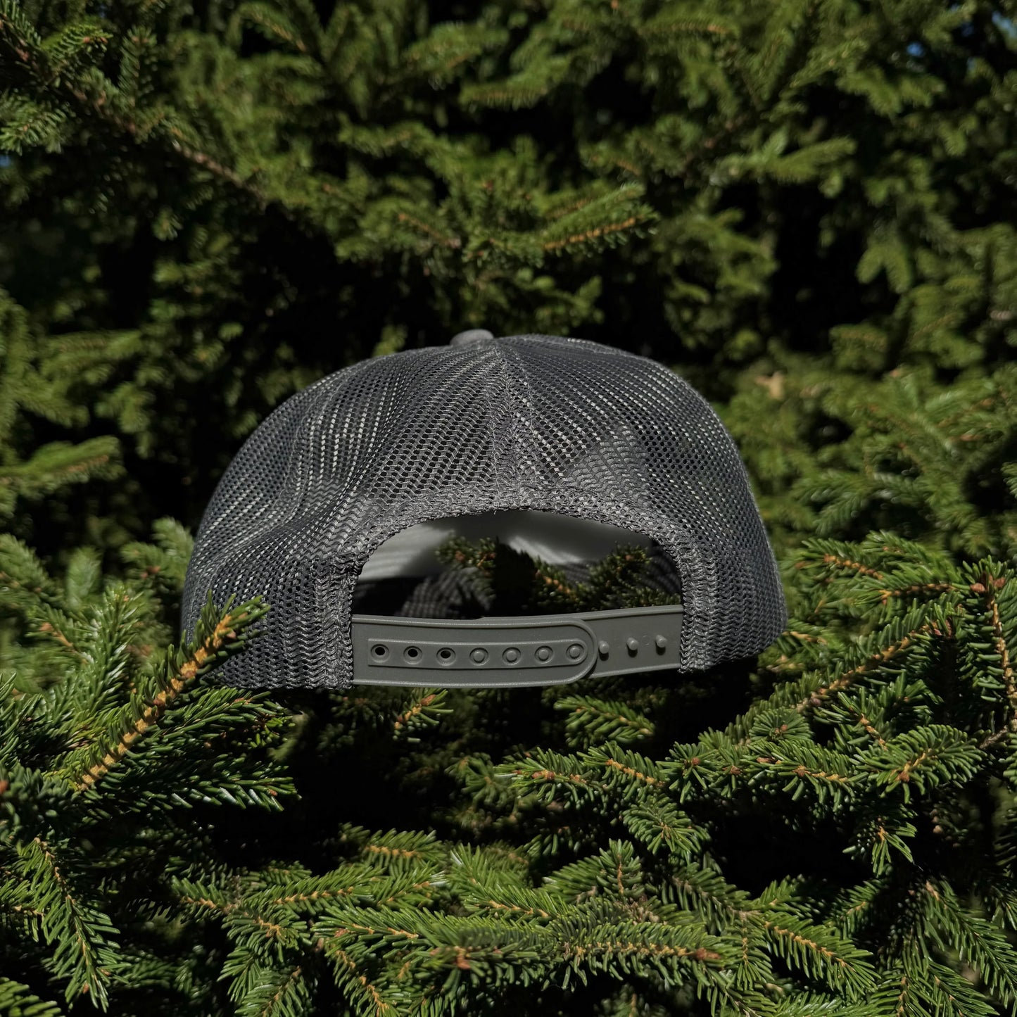 A back view of a PAKO Designs coal colored 5 panel foam trucker hat with the Tetoniac logo in Seafoam displayed against a backdrop of green pine needles.