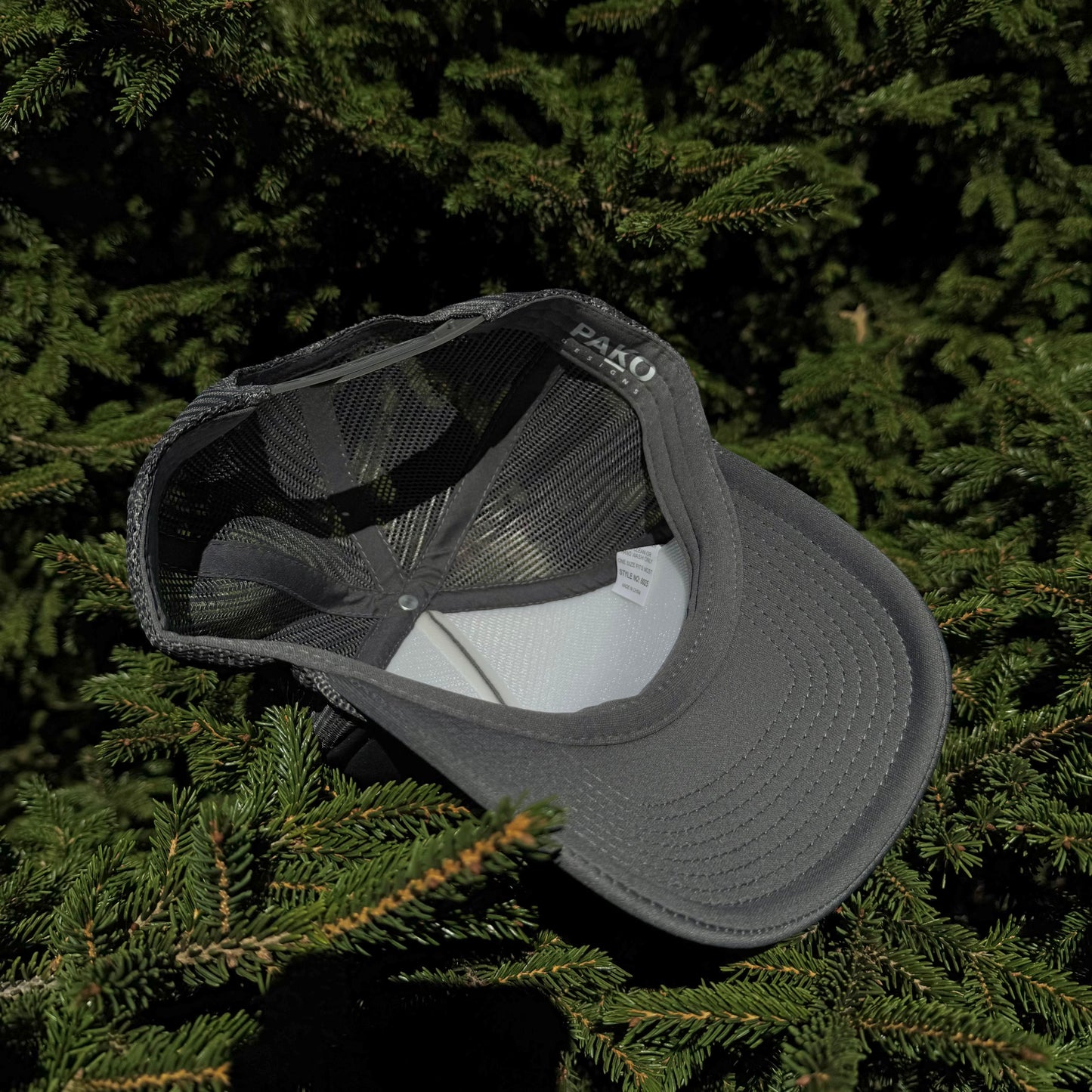 An inside view of a PAKO Designs coal colored 5 panel foam trucker hat with the Tetoniac logo in Seafoam displayed against a backdrop of green pine needles.