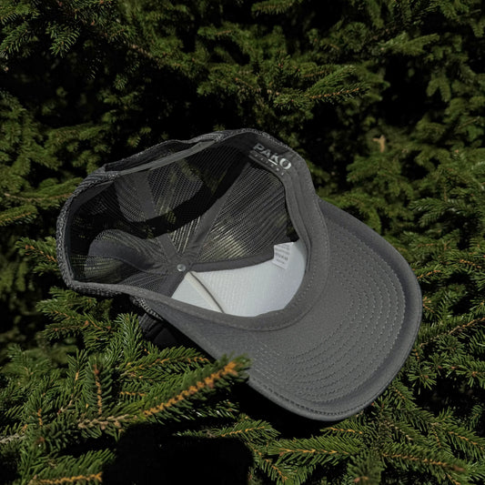 An inside view of a PAKO Designs coal colored 5 panel foam trucker hat with the Tetoniac logo in Seafoam displayed against a backdrop of green pine needles.
