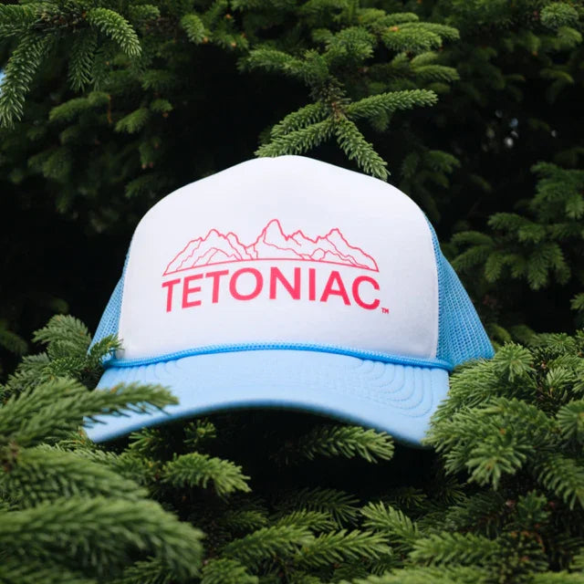A 5 panel foam trucker hat in baby blue, white, and red, with the word 'TETONIAC' printed on the front, displayed against a backdrop of a pine tree