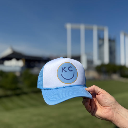 Blue and white KC Smile foam trucker hat with 'KC' logo held in front of a stadium.