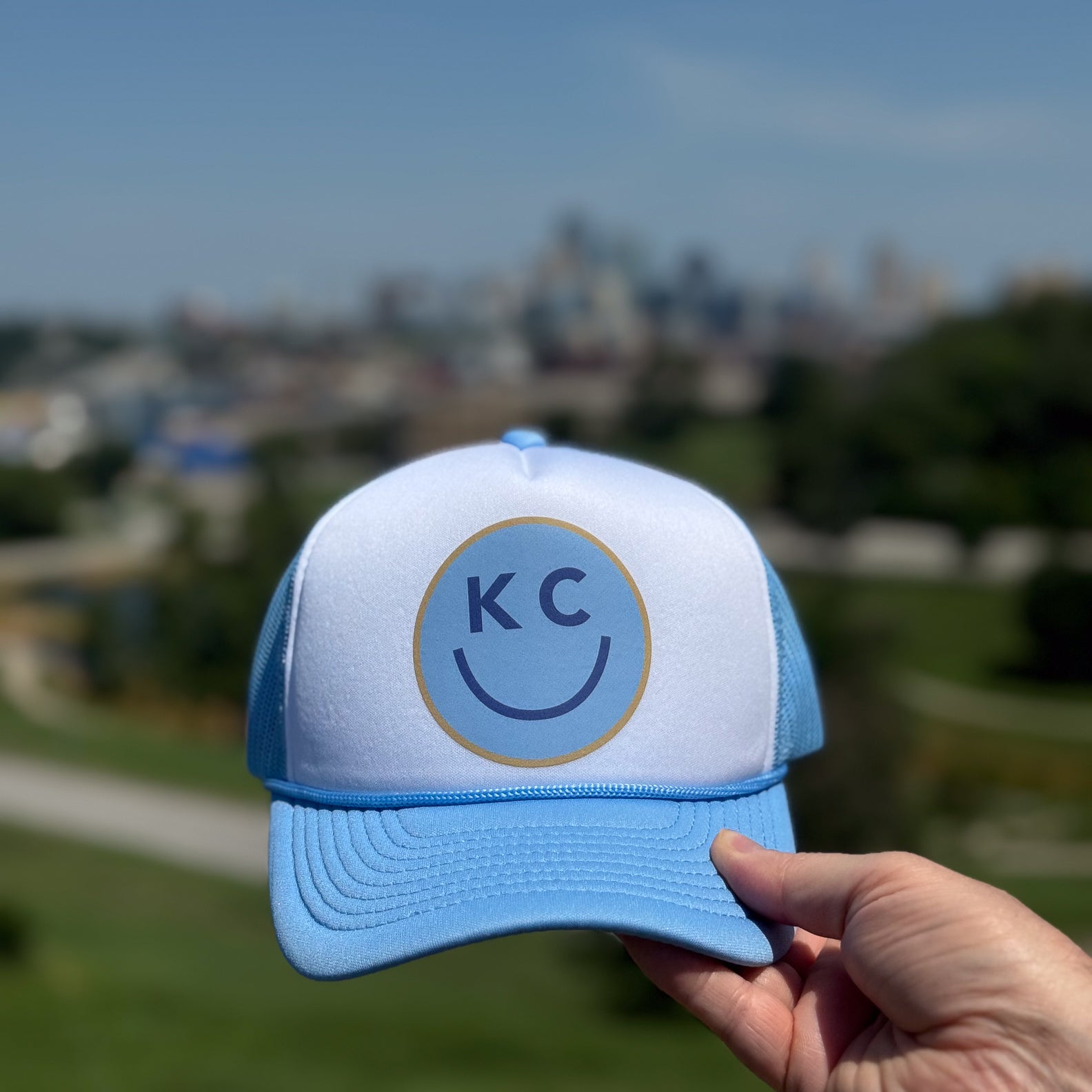 Blue and white KC Smile foam trucker hat with 'KC' logo held in front the Kansas City skyline.