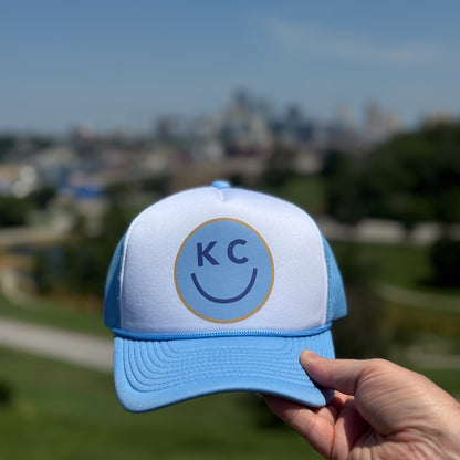 Blue and white KC Smile foam trucker hat with 'KC' logo held in front the Kansas City skyline.