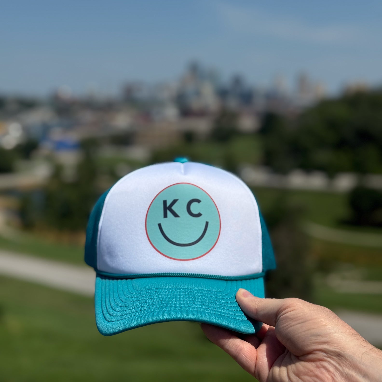 Teal and white cap with 'KC' logo held in front of the Kansas City skyline