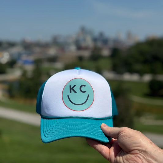 Teal and white cap with 'KC' logo held in front of the Kansas City skyline