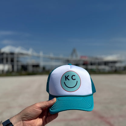 Teal and white cap with 'KC' logo held in front of a CPKC stadium
