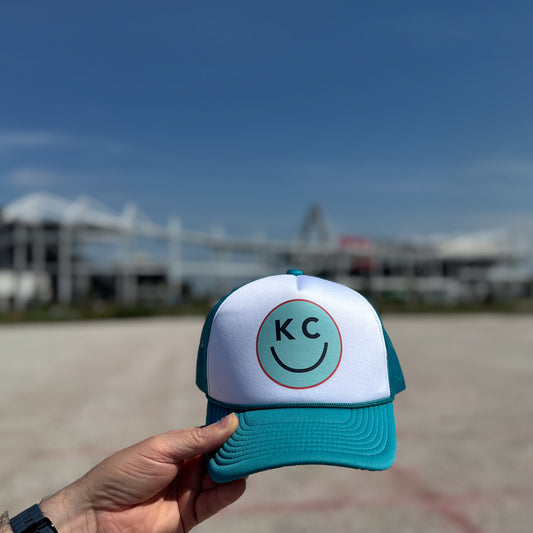 Teal and white cap with 'KC' logo held in front of a CPKC stadium