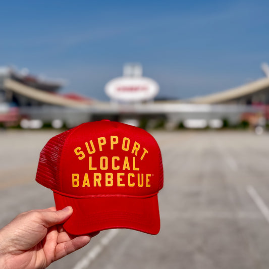Red trucker hat with 'Support Local Barbecue' text in yellow, held in front of a football stadium.