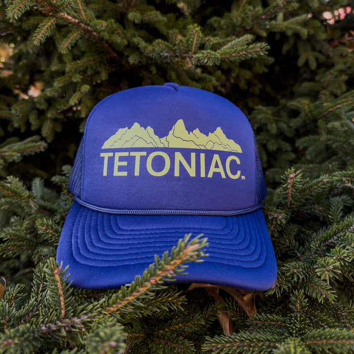 Blue cap with 'TETONIAC' text and mountain design on a green pine tree background