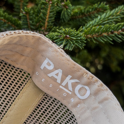 Close-up of a beige cap with 'PAKO DESIGNS' logo against a green leafy background