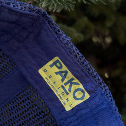 Blue fabric with a yellow 'PAKO DESIGNS' label against a blurred green background