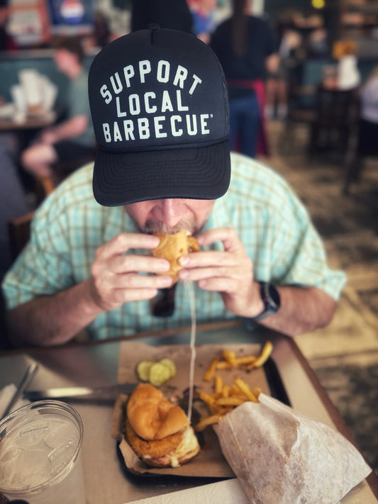 Person wearing a cap with 'Support Local Barbecue' text, eating a sandwich with a blurred background of people and food.