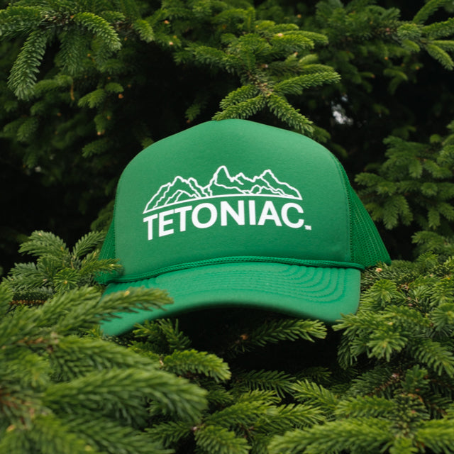 A PAKO Designs Kelly Green and White 5 panel foam trucker hat with the Tetoniac logo on the front, displayed against a backdrop of a pine tree