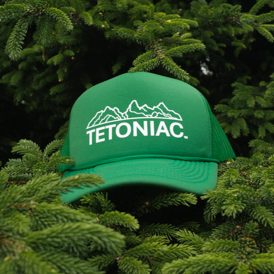 A PAKO Designs Kelly Green and White 5 panel foam trucker hat with the Tetoniac logo on the front, displayed against a backdrop of a pine tree