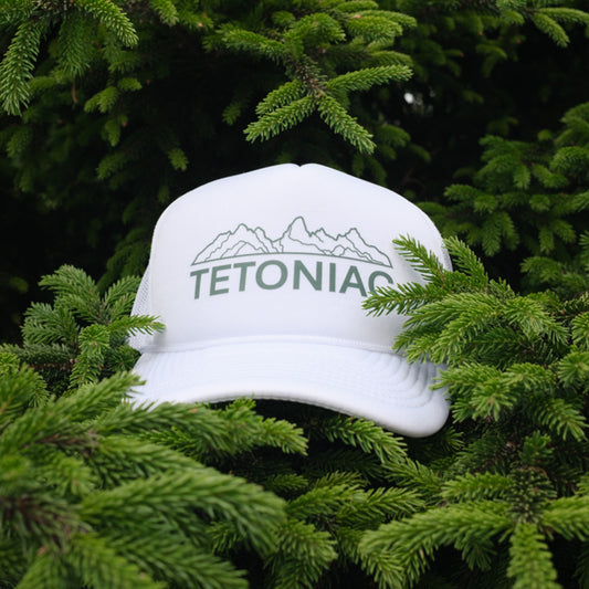 A PAKO Designs white 5 panel foam trucker hat with Tetoniac design on the front, displayed in a pine tree