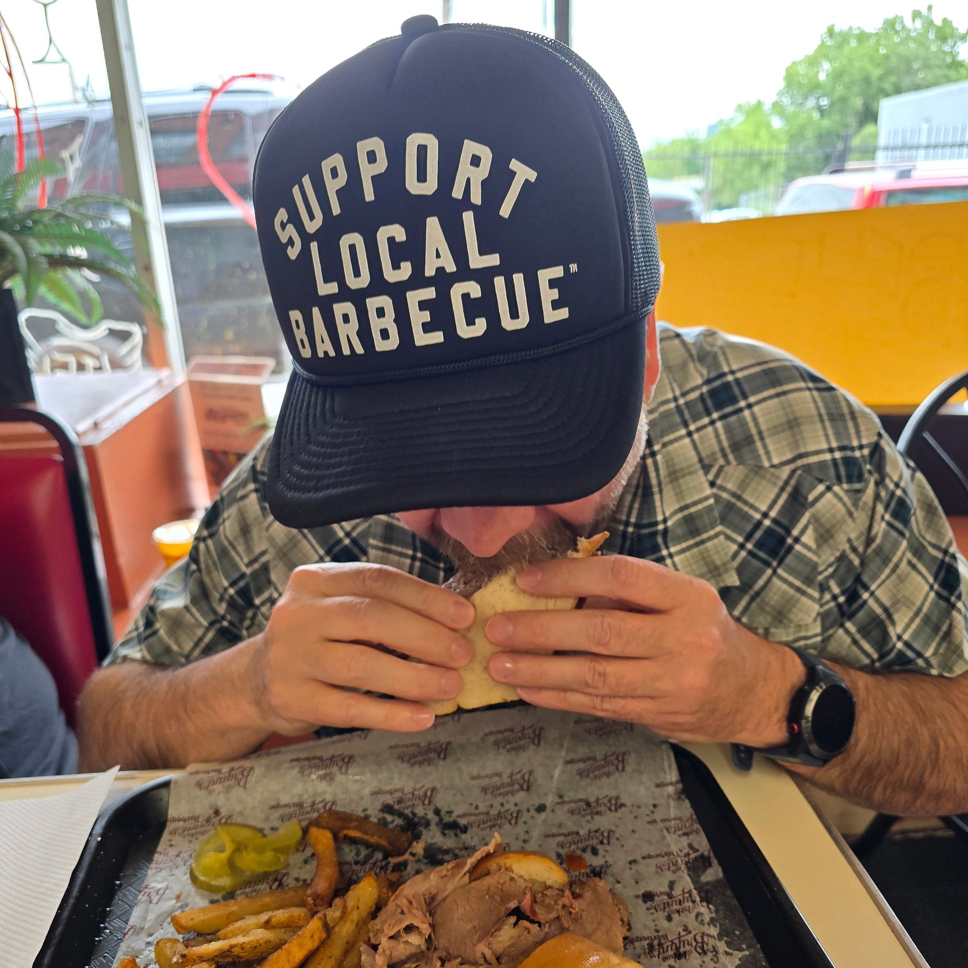 Man eating a sandwich with fries and a drink at a barbecue bbq restaurant, wearing a pako designs Support Local barbecue™ trucker hat and plaid shirt.
