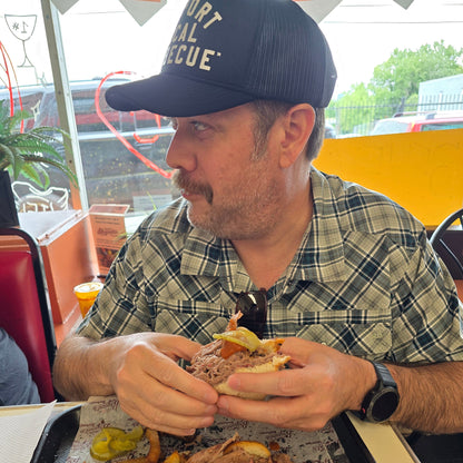 Man eating a sandwich with fries and a drink at a barbecue bbq restaurant, wearing a pako designs Support Local barbecue™ trucker hat and plaid shirt.