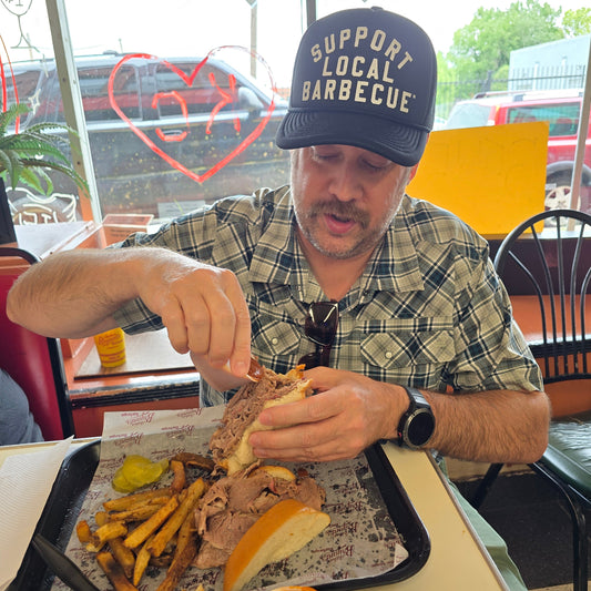 Man eating a sandwich with fries and a drink at a barbecue bbq restaurant, wearing a pako designs Support Local barbecue™ trucker hat and plaid shirt.