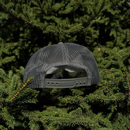 A back view of a PAKO Designs coal colored 5 panel foam trucker hat with the Tetoniac logo in Seafoam displayed against a backdrop of green pine needles.