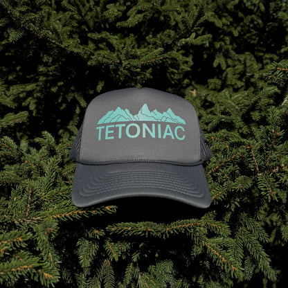 A coal colored 5 panel foam trucker hat with the Tetoniac logo in Seafoam displayed against a backdrop of green pine needles.