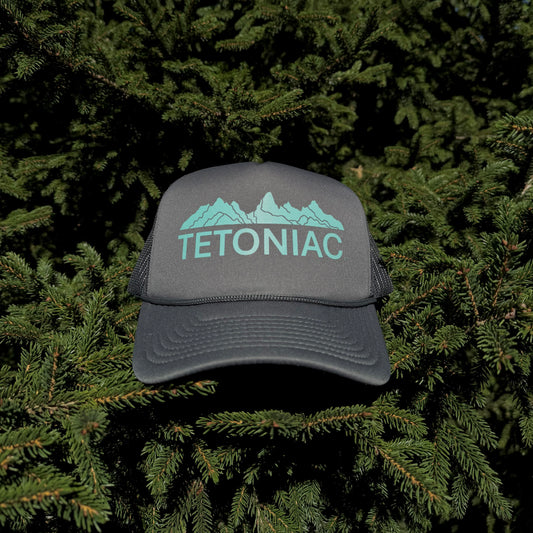 A coal colored 5 panel foam trucker hat with the Tetoniac logo in Seafoam displayed against a backdrop of green pine needles.