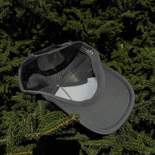 An inside view of a PAKO Designs coal colored 5 panel foam trucker hat with the Tetoniac logo in Seafoam displayed against a backdrop of green pine needles.