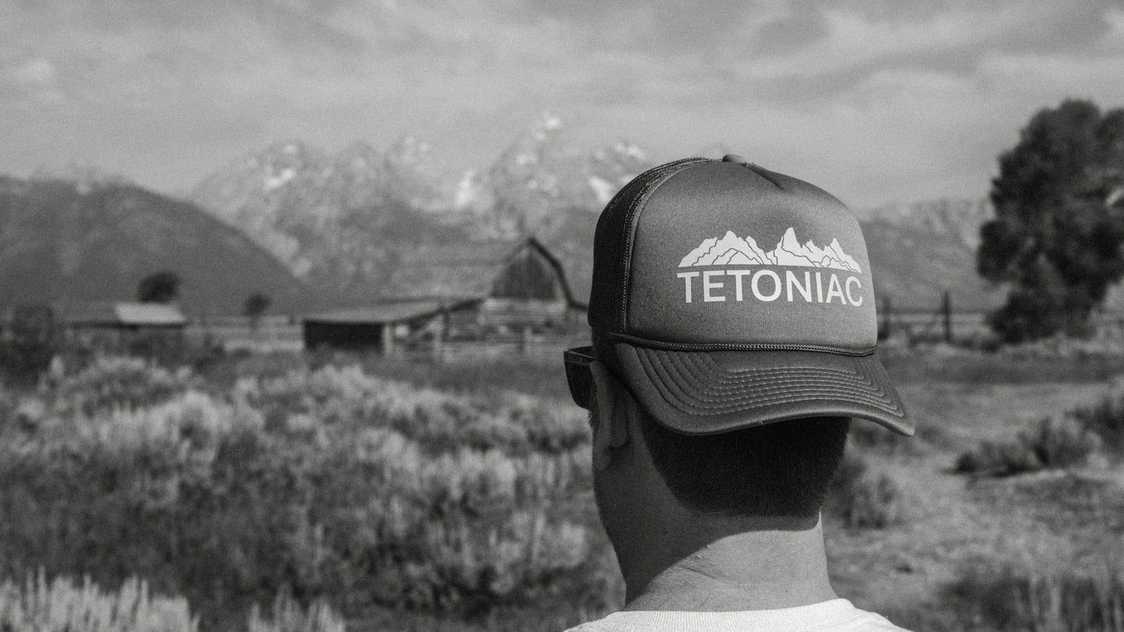 Person wearing a 'Tetoniac' cap with Teton Mountains and Moulton Barn in the background