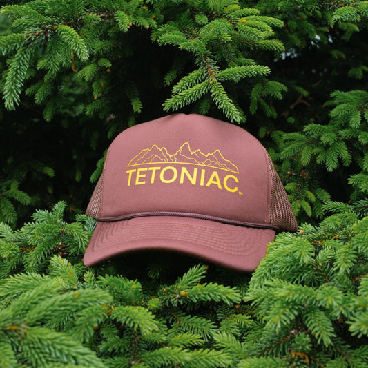 A brown and yellow 5 panel foam trucker hat with the word 'TETONIAC' printed on the front, displayed against a backdrop of green foliage.