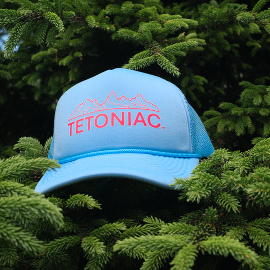 A baby blue and red Tetoniac 5 panel foam trucker hat displayed in a pine tree