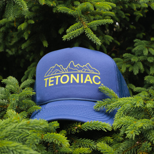 A navy blue 5 panel foam trucker hat with the Tetoniac logo in PAKO Green displayed against a backdrop of green pine needles.
