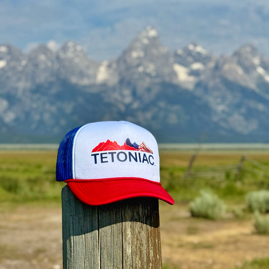 PAKO Designs Red,White & Blue Trucker hat with Stars & Stripes 'TETONIAC' logo on a wooden post with the Teton Mountains in the background